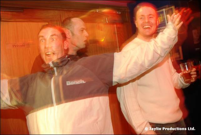 Two people with arms raised and mouths open cheer in a club, one in a Bench jacket holding a drink