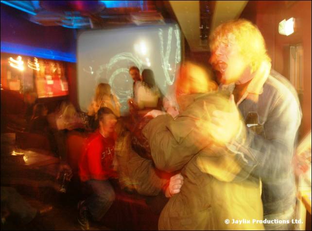 A blurred dance scene with a person in a green coat and another with blonde hair in the foreground
