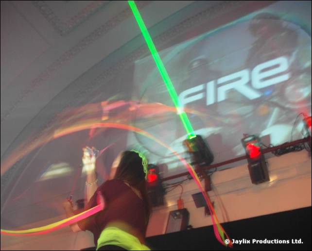 A dancer with a glowing hoop stands before a projected FIRE logo and a green laser beam