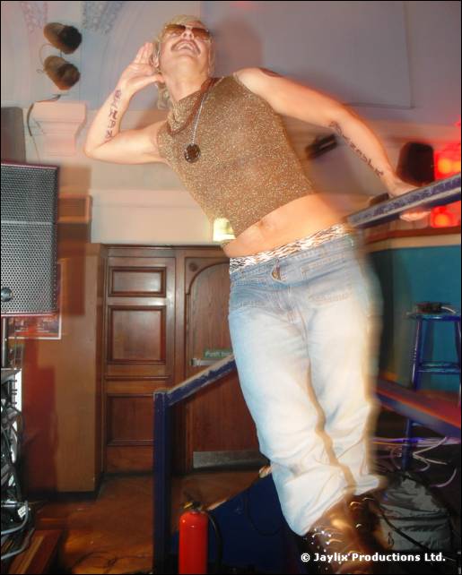 A person in a glittery gold top and light jeans dances on a raised platform, one hand behind their head