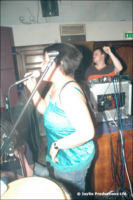 A person sings into a microphone in the foreground while a DJ at the decks raises an arm behind