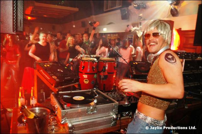 A person in a blonde wig and sunglasses stands at DJ decks with congas and a dancing crowd behind