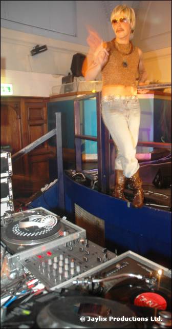 A person in a blonde wig and glittery top stands on a raised platform above DJ turntables