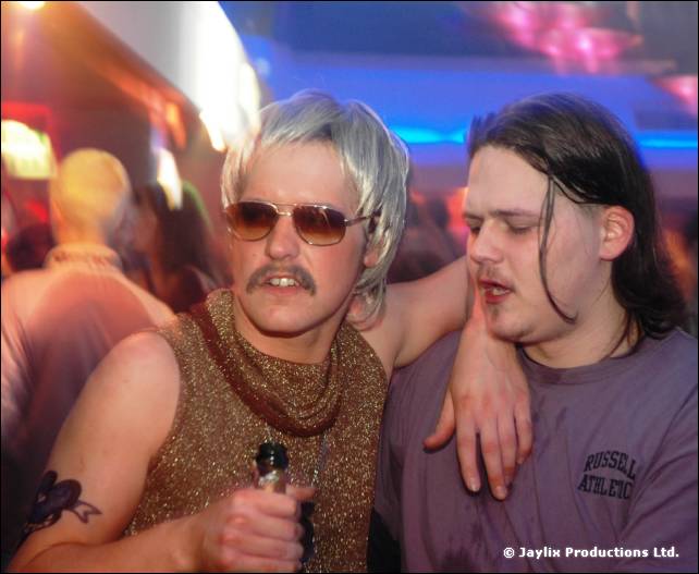 Two people stand with arms round each other, one in a blonde wig, sunglasses and glittery top