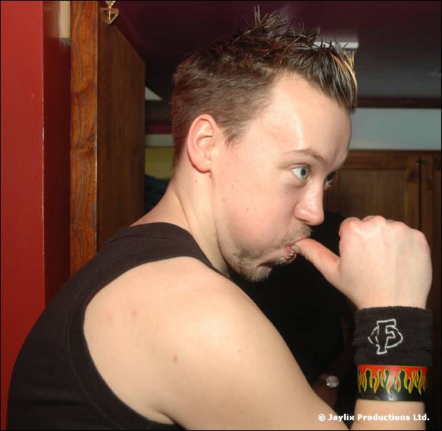 A person in a black vest bites a knuckle and looks sideways, wearing a flame patterned wristband