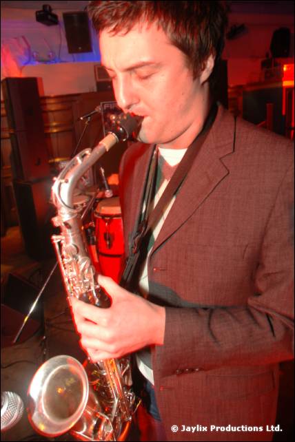 A person in a brown jacket plays a saxophone with eyes down, speakers and a conga behind