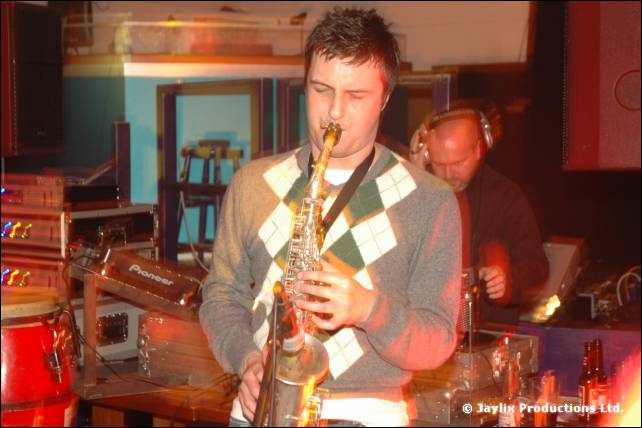 A person in a diamond patterned jumper plays saxophone, with a DJ in headphones behind