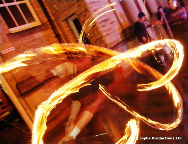 A person spins loops of fire in front of a brick building, the figure blurred behind the flames