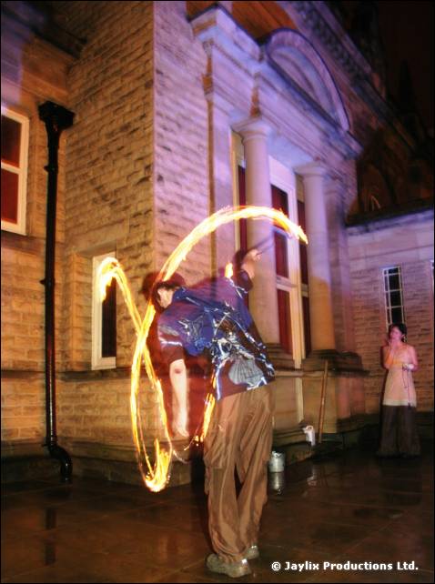 A person in baggy trousers spins fire poi beside a stone building, another person standing behind
