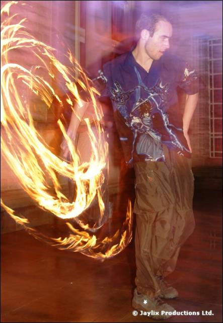 A person in a dark patterned shirt and baggy trousers stands with fire poi trailing behind them