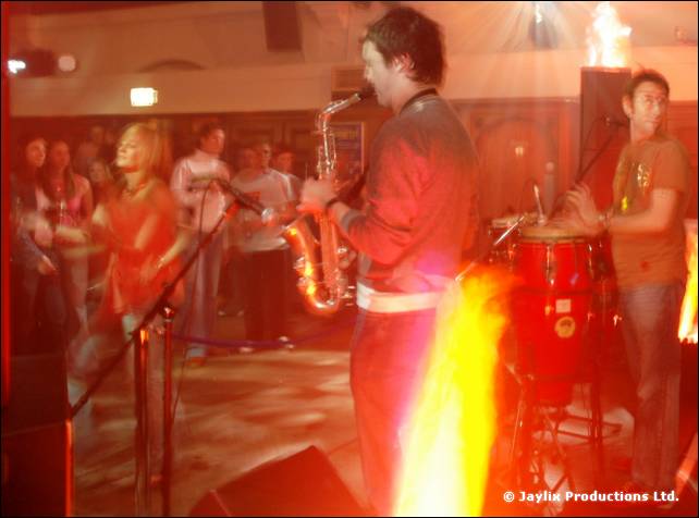 A saxophonist and a conga player perform on stage with flames in the foreground and a crowd behind