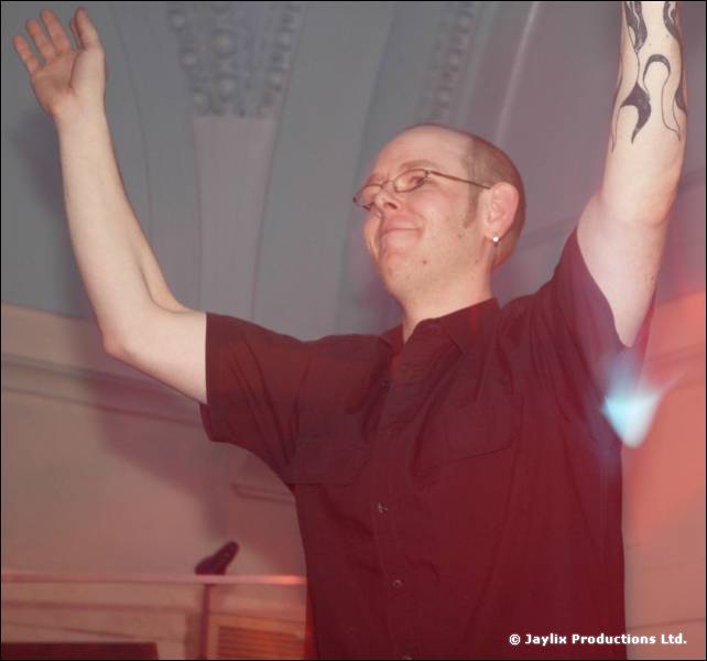 A man with a shaved head and glasses raises both arms in a hall with an ornate ceiling, a flame tattoo on one arm