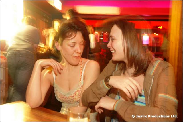 Two people laugh together at a bar table, one in a vest top and one in a hooded cardigan