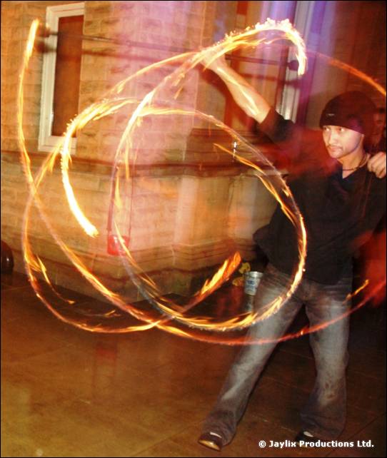 A person in a black hat and top spins fire poi near a brick wall at night