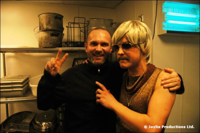 A smiling man in a black jumper poses with a person in a blonde wig and sunglasses in a kitchen, making a peace sign