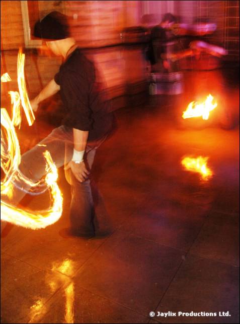 A person in a dark hat spins fire at night, blurred, with small flames on the floor behind
