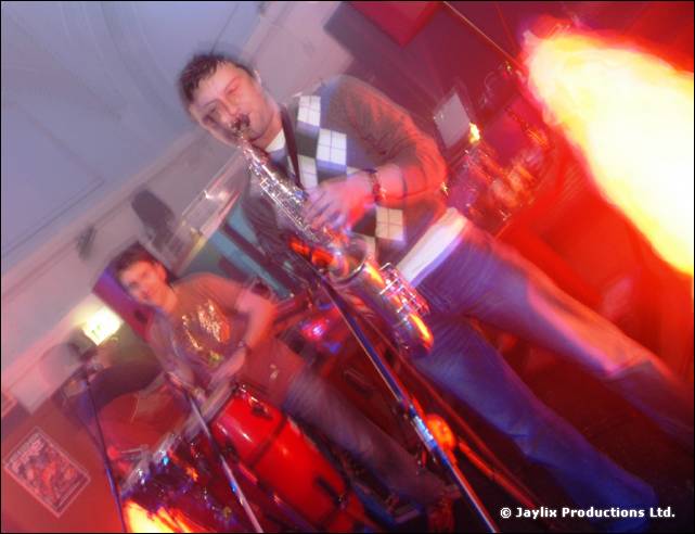 A saxophonist in a diamond patterned jumper plays in front of a flame effect, tilted view