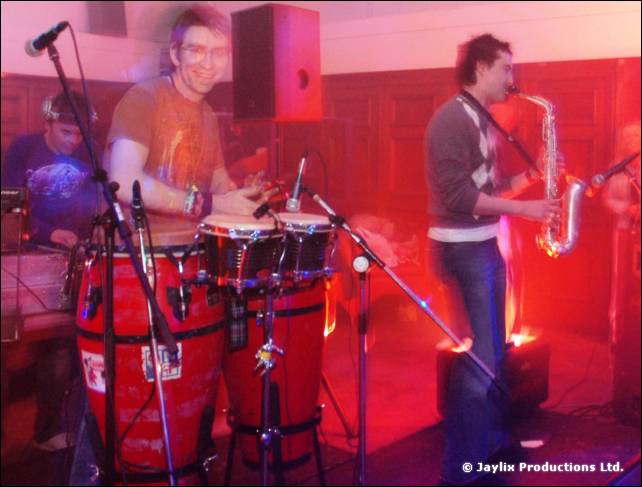 A person plays congas and another plays saxophone on a stage lit in pink and red
