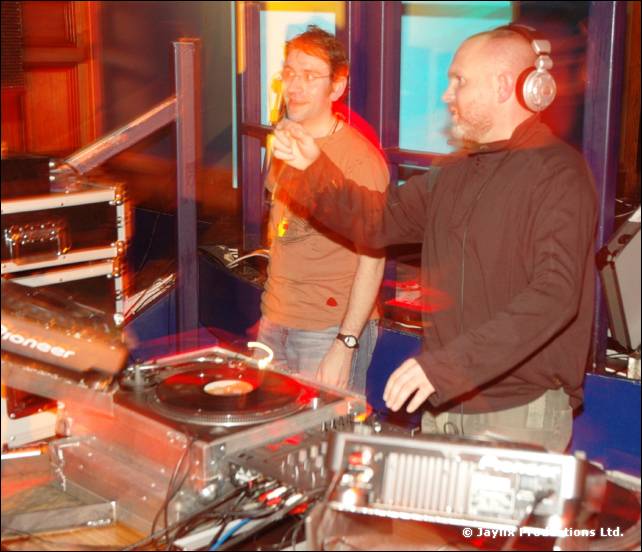 Two people at DJ decks, one in a brown top and headphones, the other holding a microphone