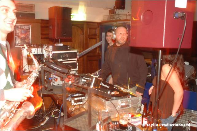 A saxophonist at left, with a DJ in headphones behind a table of decks and equipment