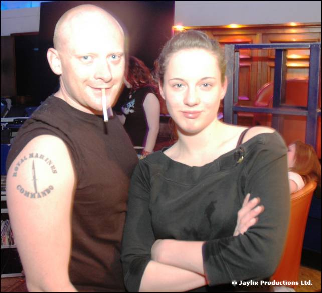 Two people pose for the camera, one bald with a cigarette in his mouth and a Royal Marines tattoo
