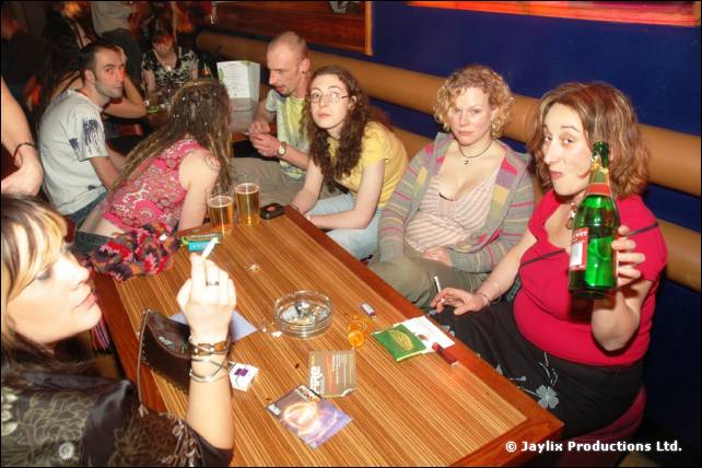 People sit around a wooden table with drinks, an ashtray and cigarettes, one holding a green bottle