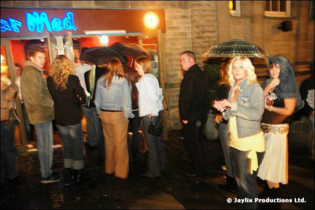 People queue in the rain outside Bar Med at night, some holding umbrellas