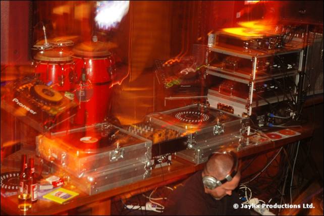 DJ decks, a mixer and congas on a table, with a person in headphones crouched at the bottom
