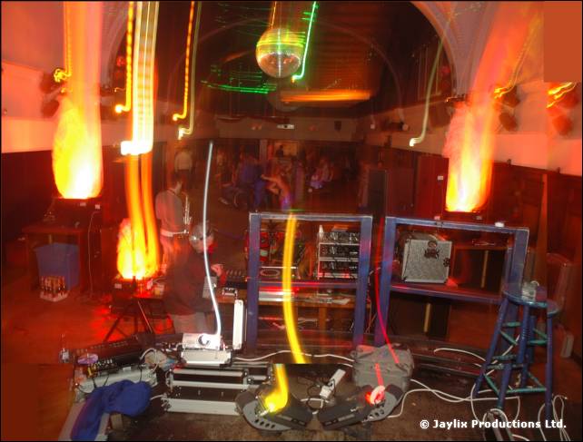 A hall with flame effects, a mirror ball and DJ equipment on the floor, lit with long light trails