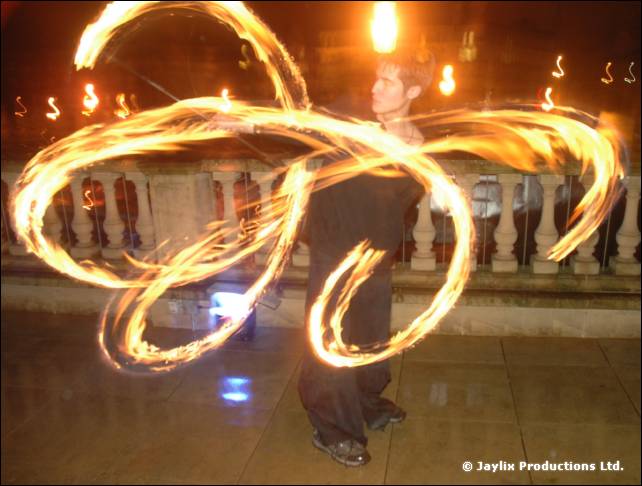 A person spins fire poi in large loops on a terrace with a stone balustrade behind