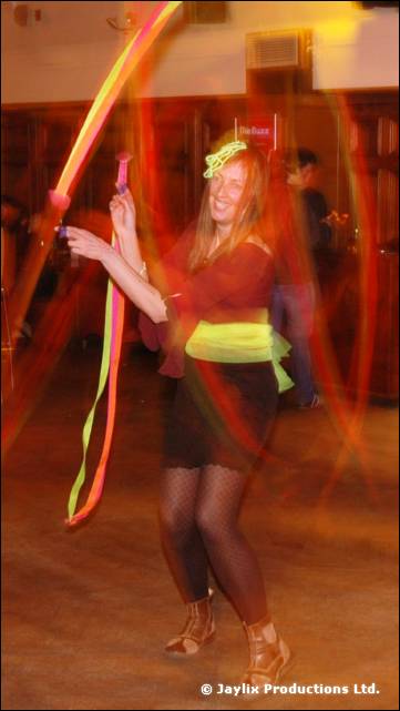 A smiling person with long hair dances holding coloured ribbons, in a yellow green waist wrap
