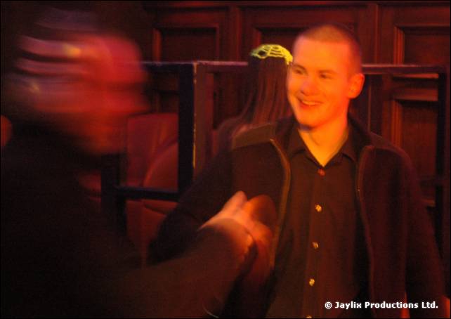 A smiling person with cropped hair in a dark shirt, with a blurred figure in the foreground