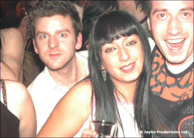 Three people pose close together at the camera, one holding a glass, in a bright flash