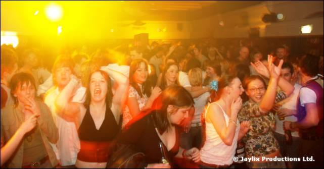 A crowd on a dance floor under yellow light, people with hands raised and mouths open