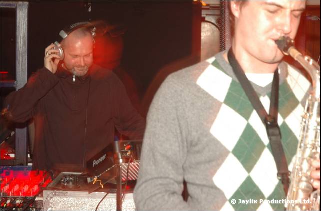 A DJ in headphones at Pioneer decks stands behind a saxophonist in a diamond patterned jumper