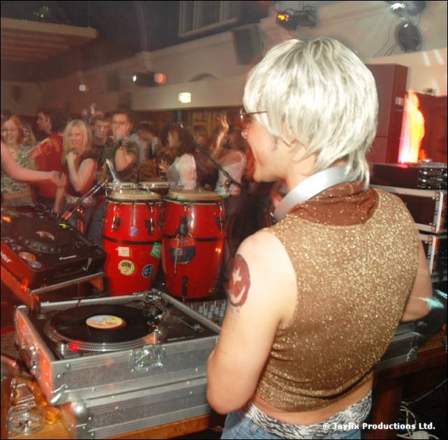 A person in a blonde wig and glittery top stands at DJ decks, seen from behind, with congas