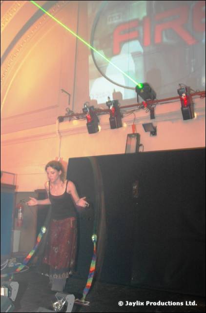 A person in black stands on a stage below a projected FIRE logo, a green laser and stage lights