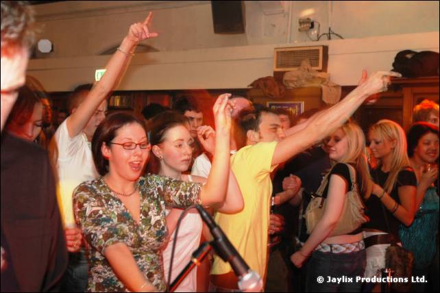 People on a crowded dance floor with arms raised, one in a floral top and glasses, a microphone nearby