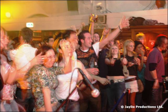 A crowd dances close to a microphone stand, arms up, in a hall with wood panelling
