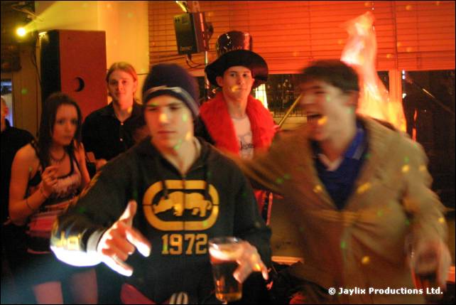 Dancers in a bar, one in a hoodie reading 1972 holding a drink, another in a dark hat
