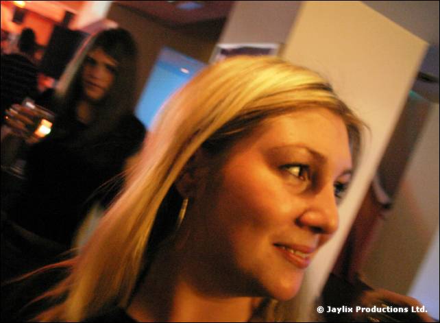 A woman with long blonde hair smiles and looks to one side in a dim bar