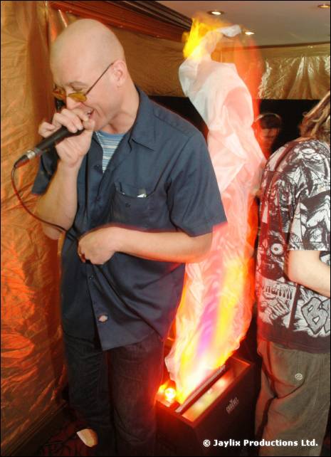 A man in sunglasses and a blue shirt sings into a microphone beside a fire effect