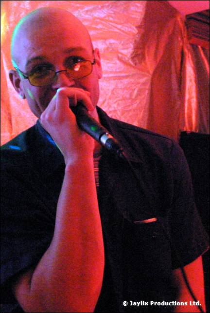 A man in tinted glasses sings into a microphone against a pink backdrop