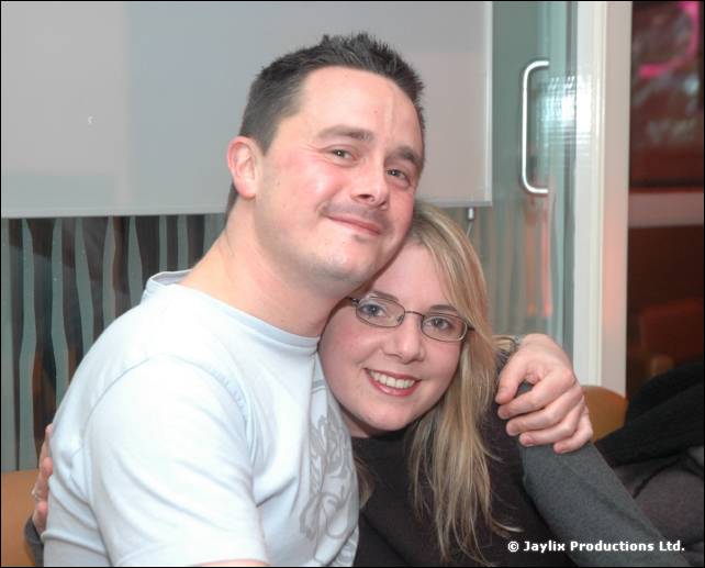 A man in a white T-shirt hugs a smiling woman in glasses