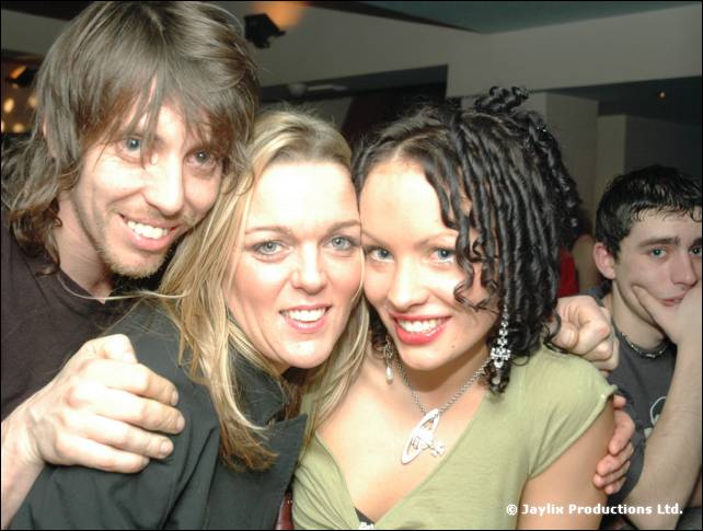 Two smiling women and a man with shoulder-length hair pose together, a young man behind them