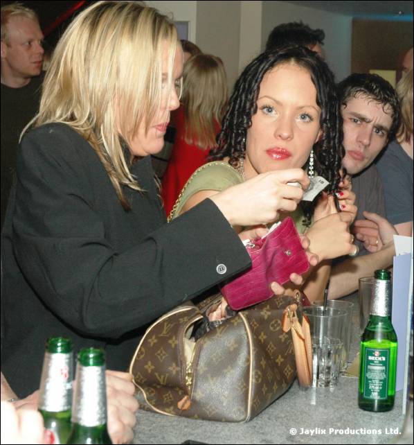 A woman in a black jacket gets money from a pink purse beside a woman with braided hair, bottles in front