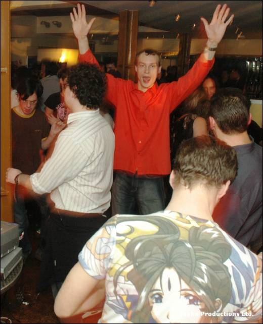 A man in a red shirt raises both arms among dancers, a person in a manga print T-shirt in front