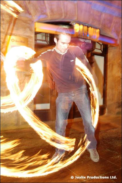 A man in a dark top swings fire in wide trails outside a stone building