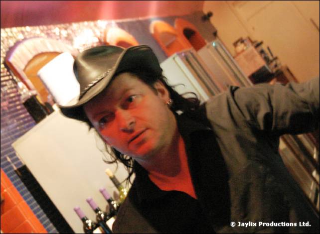 A man with long dark hair in a black cowboy hat looks at the camera in a bar