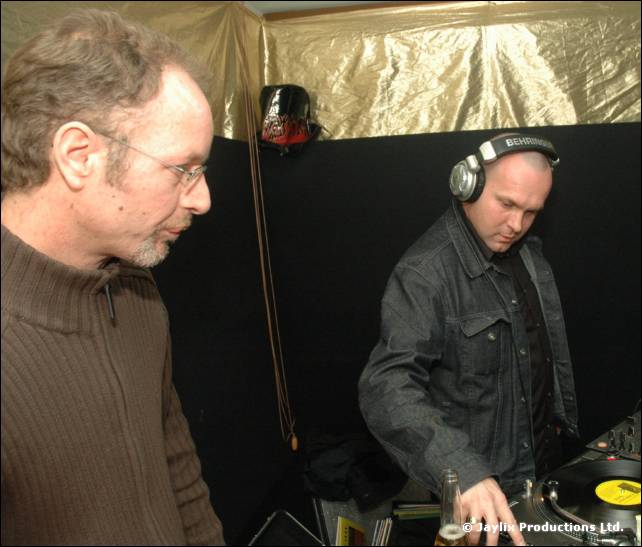 Two DJs at decks, one in a grey jumper and glasses, the other in a denim jacket and headphones
